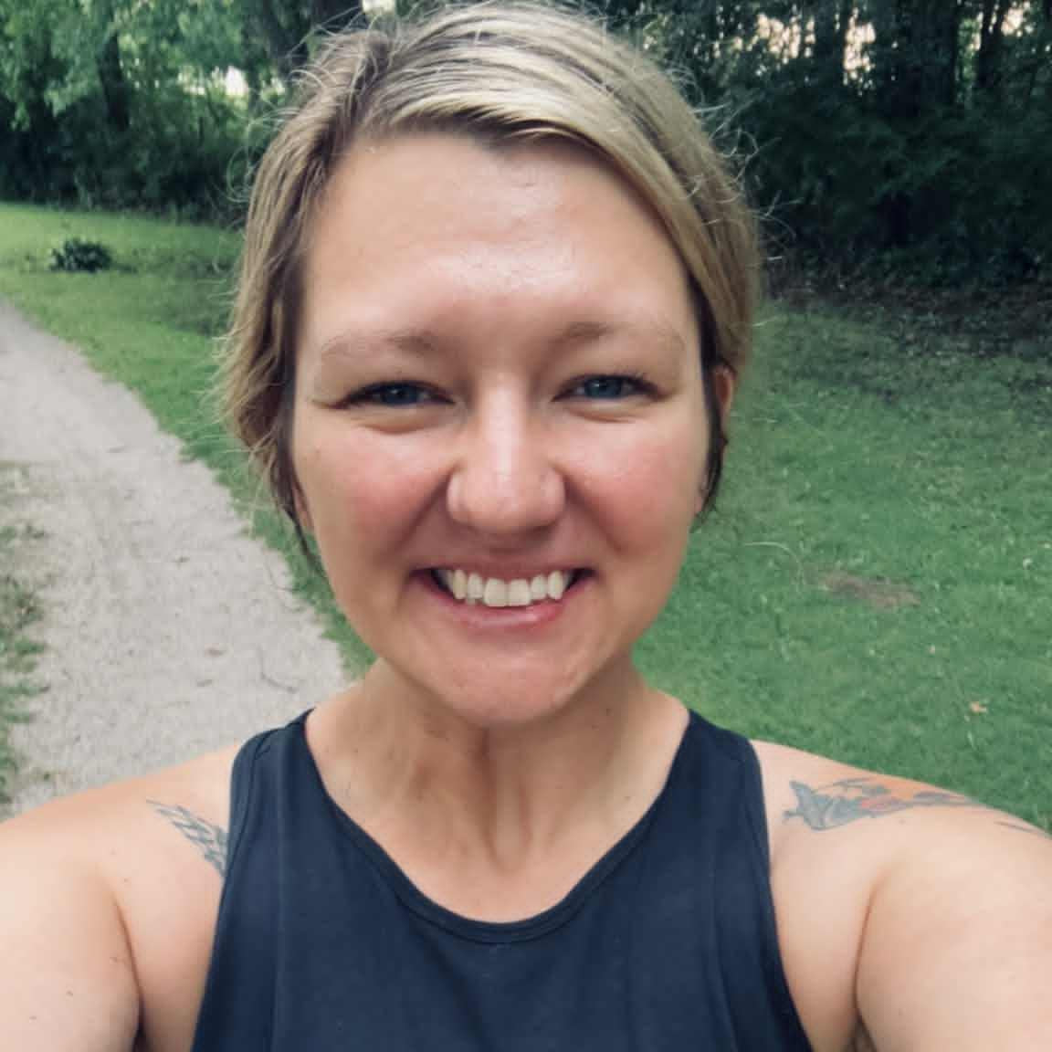 Woman in a black sleeveless top standing outdoors on a path with greenery.