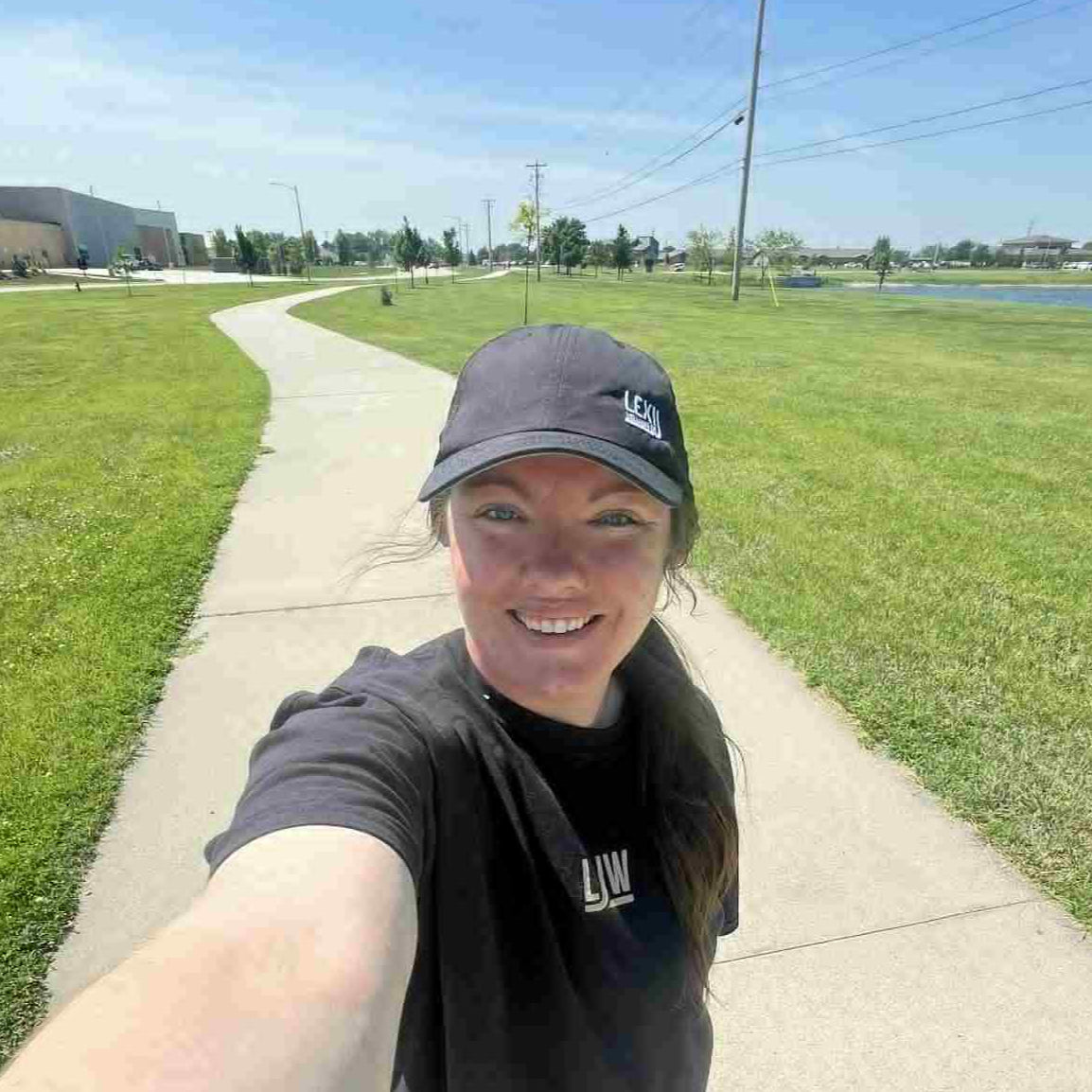 Person taking a selfie on a sidewalk with grass and buildings in the background